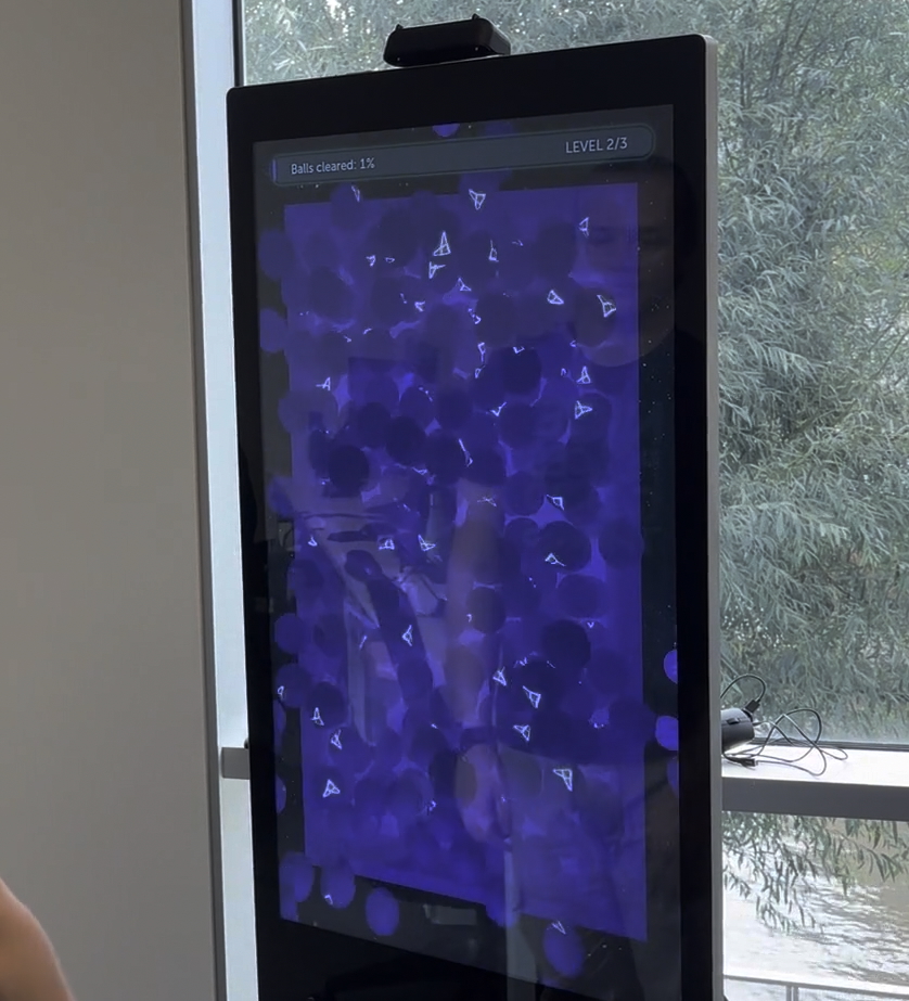 A kiosk with a brand experience application being used. The user is prompted to "clear the balls" then balls enter the screen, the user moves them around, emptying the pit. A message reading "TouchFree is engaging" is displayed under the balls.