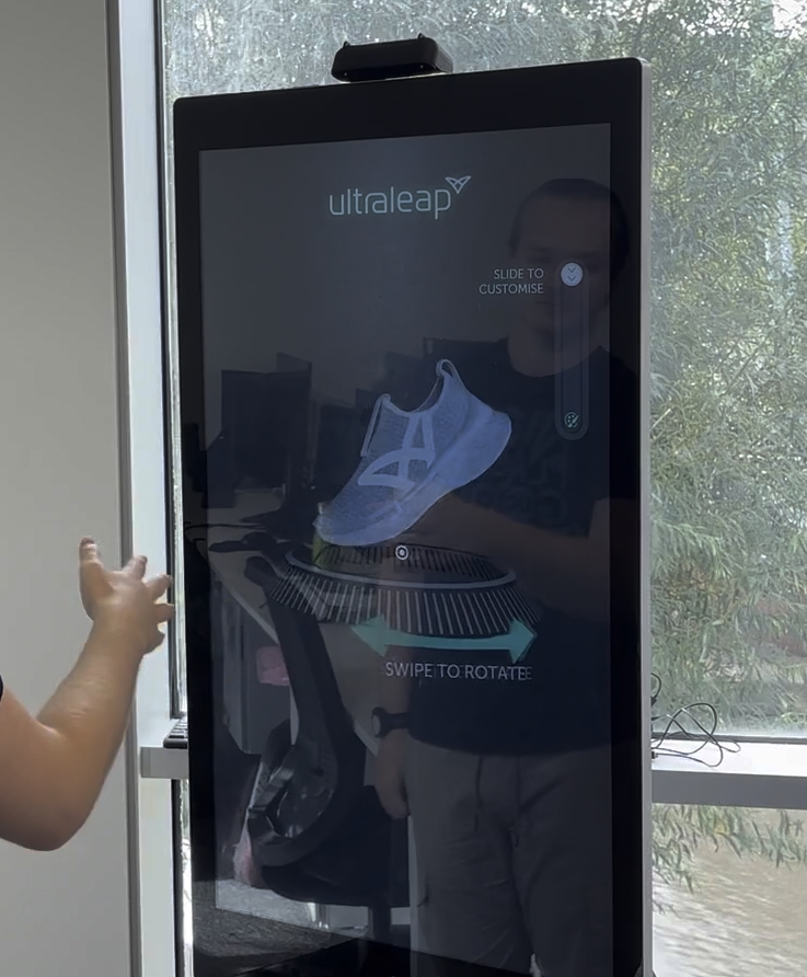 A kiosk with a product customisation application being used. The user is prompted to "pull scanner down" which reveals a training shoe. The shoe is rotated by the user swiping their hand. The shoe is expanded when a slider is moved along a track, then customised by selecting a section of the shoe and selecting a colour.